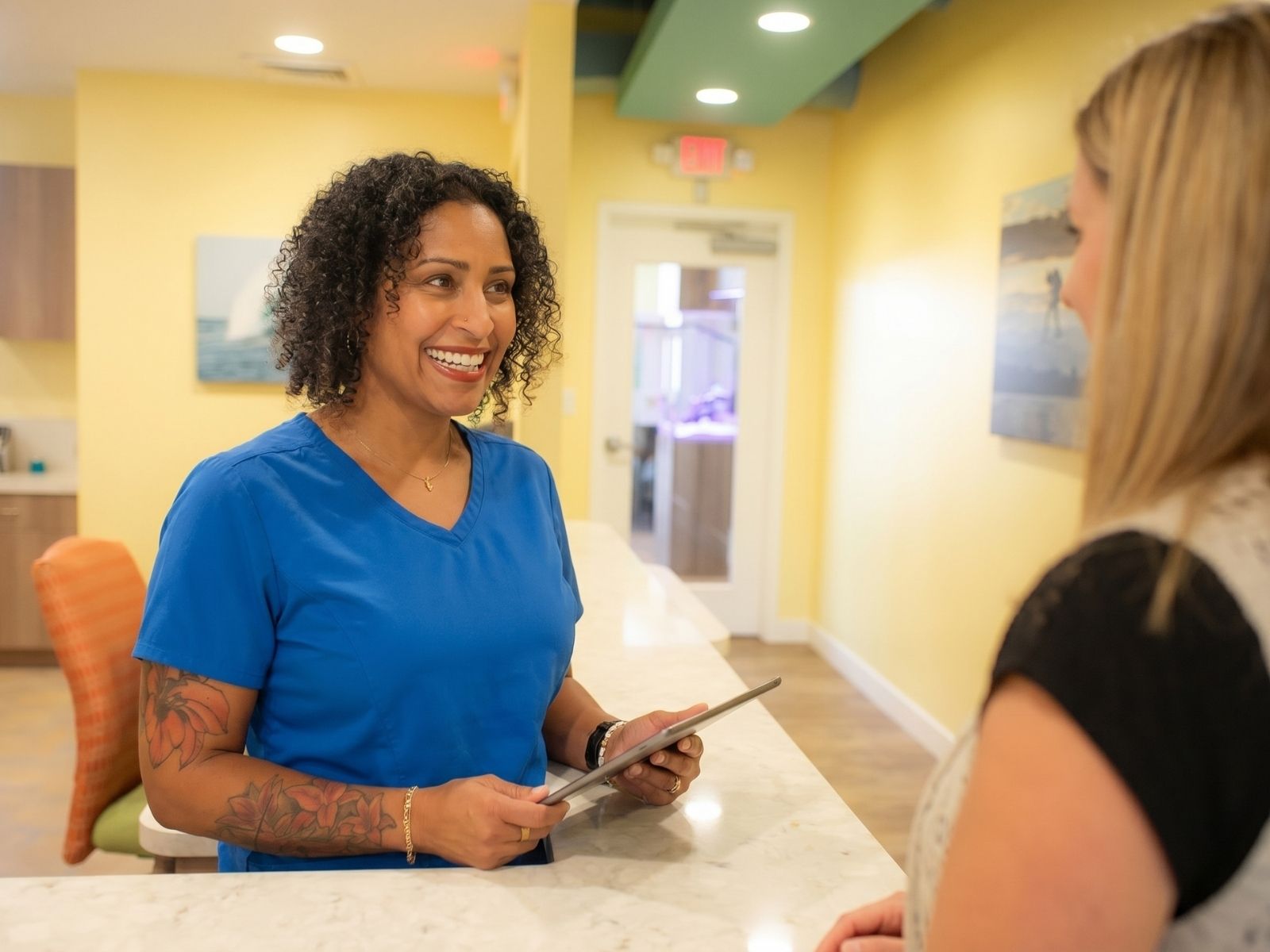 Smiling healthcare worker in blue scrubs hands a tablet to a patient at a marble reception counter in a bright clinic lobby.